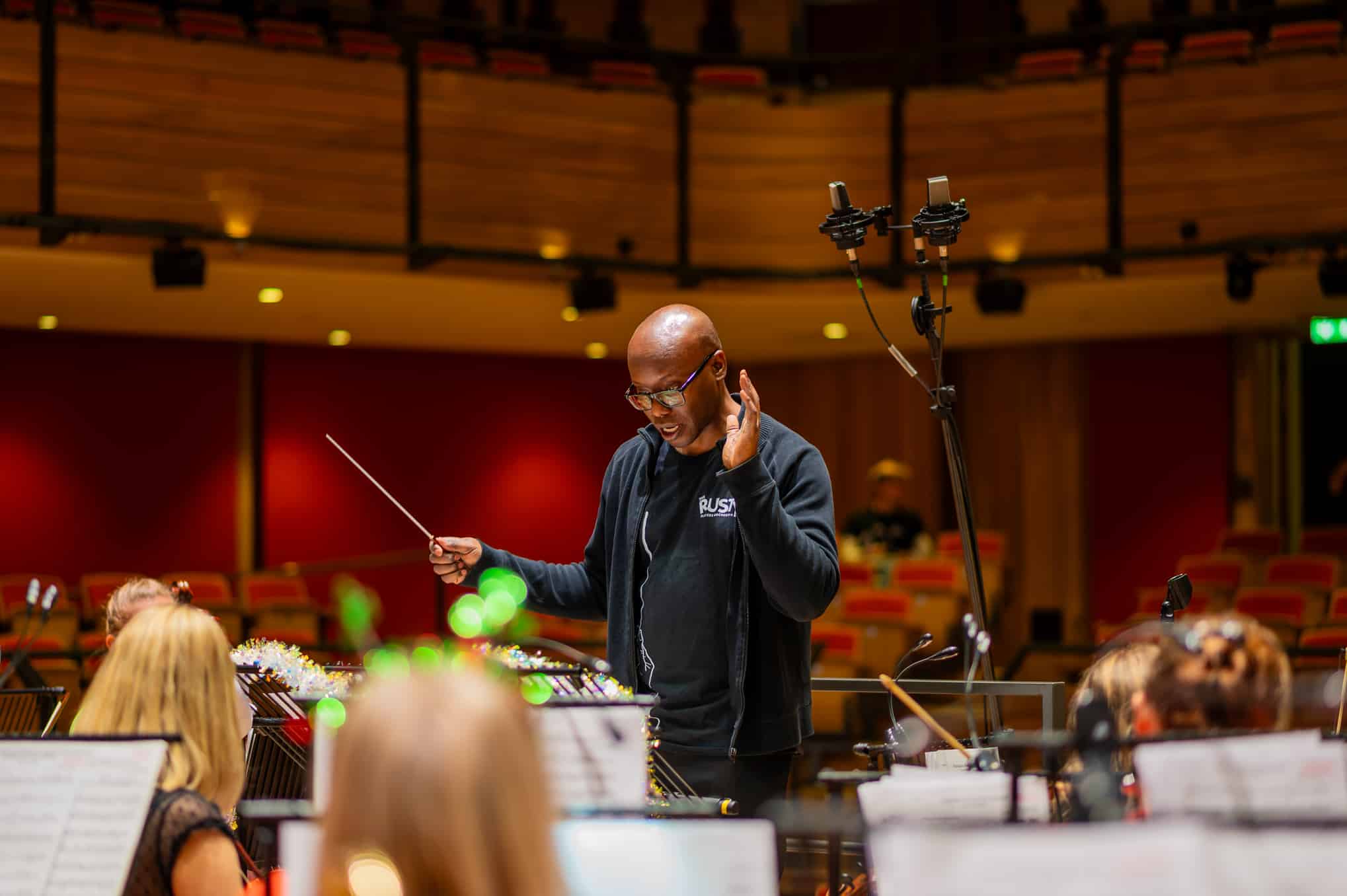 Julian Powell, wearing a black Rusty Player's Orchestra branded t-shirt, conducting the orchestra inside The Elgar Concert Hall in Birmingham.