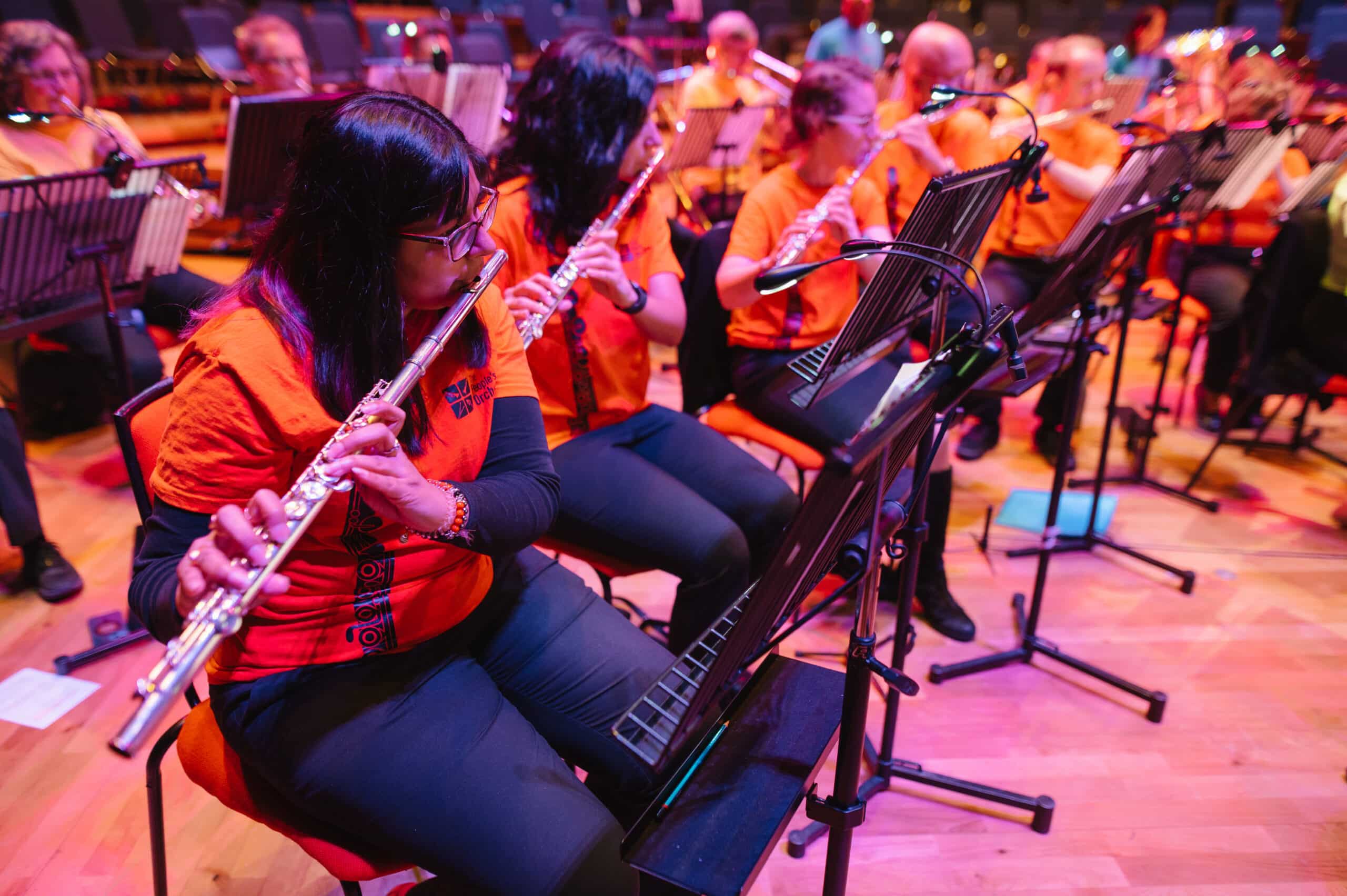 A row of flutists wearing orange t-shirts with The People's Orchestra.