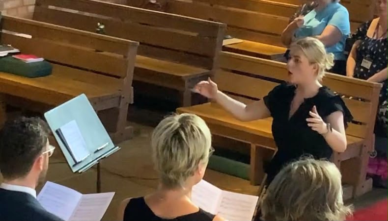 Any McCulloch, wearing a black top and tied blonde hair, is conducting a community choir in a church.