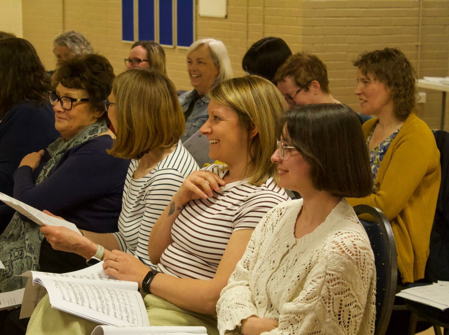 Singers in The People's Show Choir York sat in a rehearsal, smiling and laughing. 