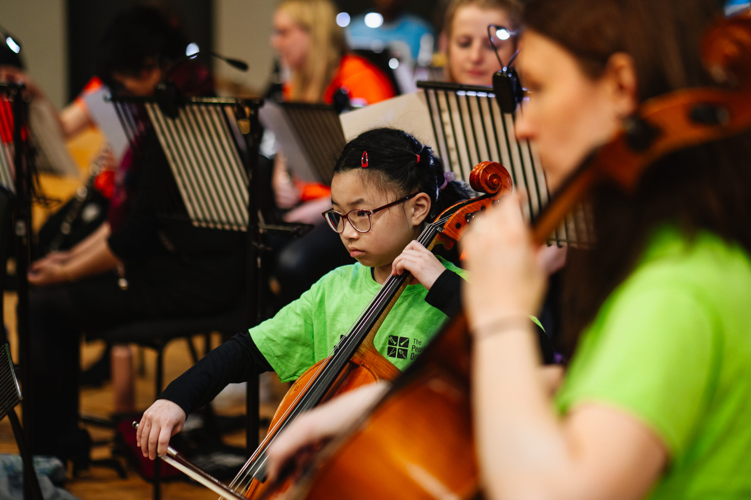 Aurora Chin-Chan is playing her cello with other musicians in The People's Orchestra. She is wearing a green TPO-branded t-shirt.