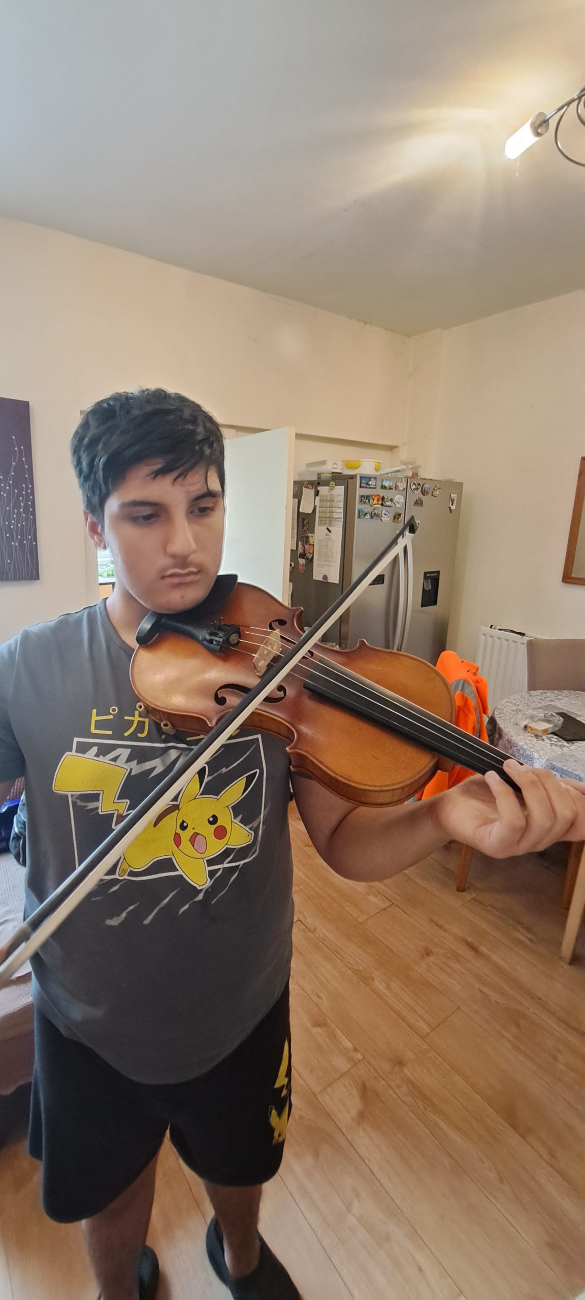 Aman Gill, who has black short hair wearing a grey t-shirt and shorts, practices violin in a living room.