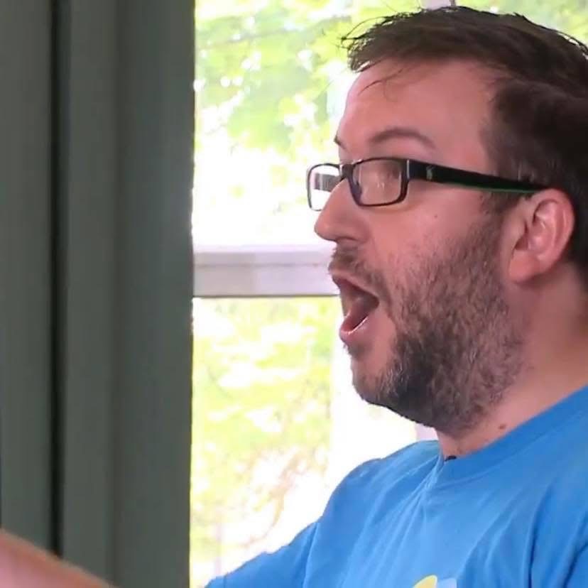 Conductor for TPSC Coventry, Andrew Johnson, conducting his choir. He wears black rimmed glasses and a blue t-shirt.