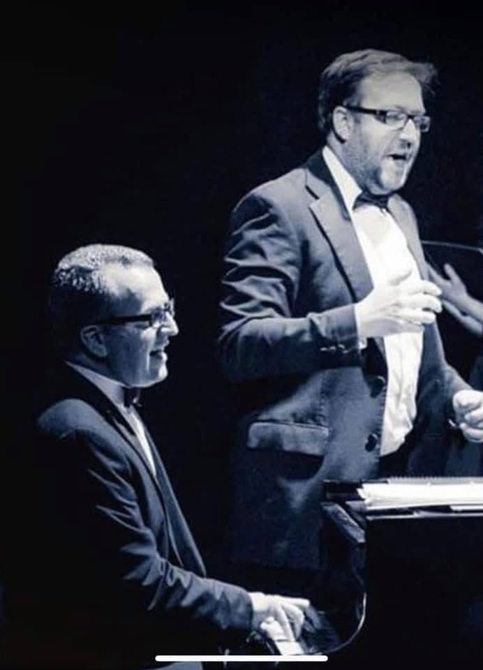 Andrew Johnson is conducting on stage with a pianist. Both men are wearing tuxedos. 