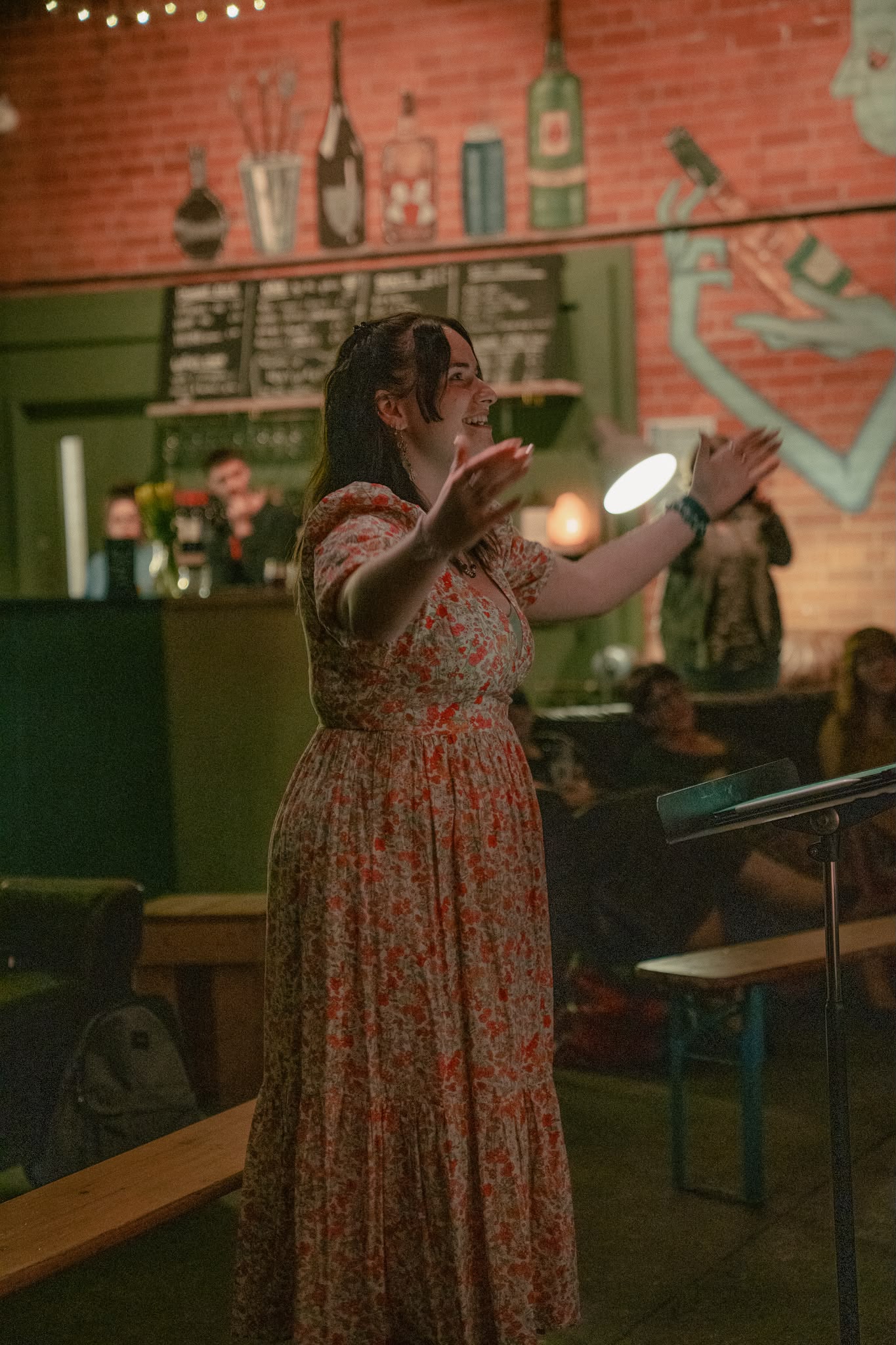 Elizabeth Cunningham, a young woman with long brown hair and wearing a red flower patterned dress, stands in a bar room conducting a choir in front of a small audience.
