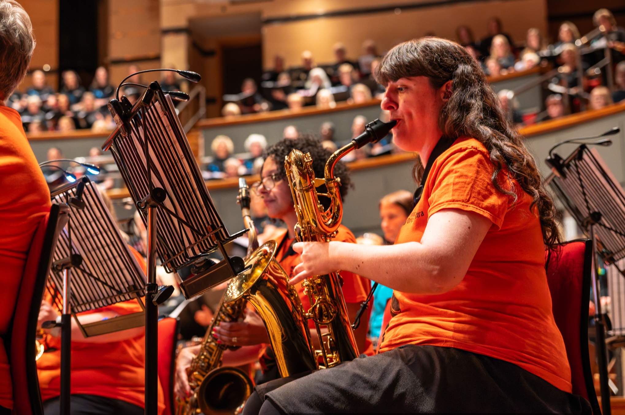 Rosie Price performs her baritone saxophone in The People's Orchestra at Birmingham Symphony Hall. She's wearing an orange People's Orchestra branded t-shirt and black trousers.