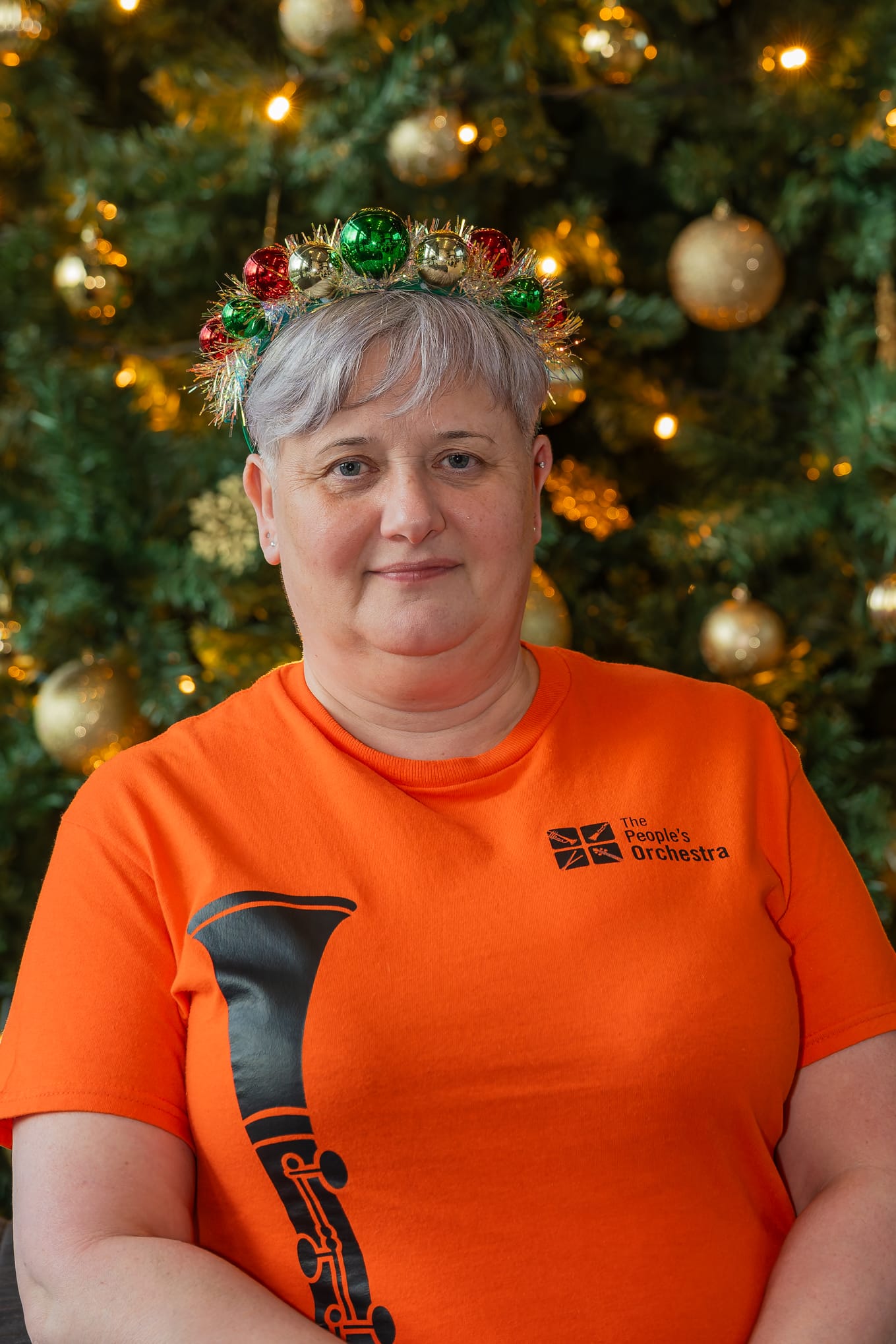 Deb Clarke stands in front of a Christmas tree. She is wearing a Christmas-themed headwear and an orange TPO-themed t-shirt.