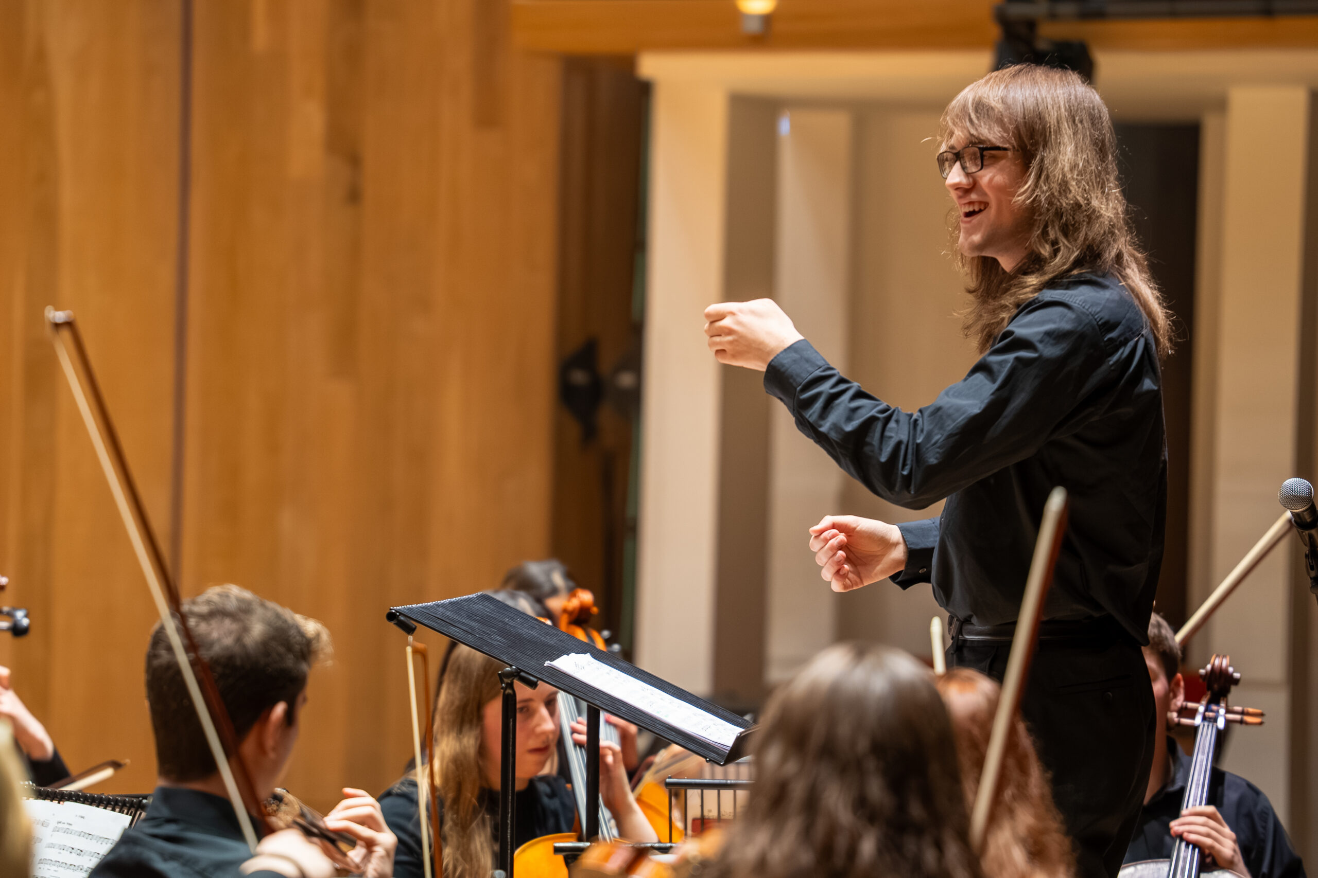 Fern Warwick, wearing a black shirt, long brown hair and black rimmed spectacles, conducts an orchestra.