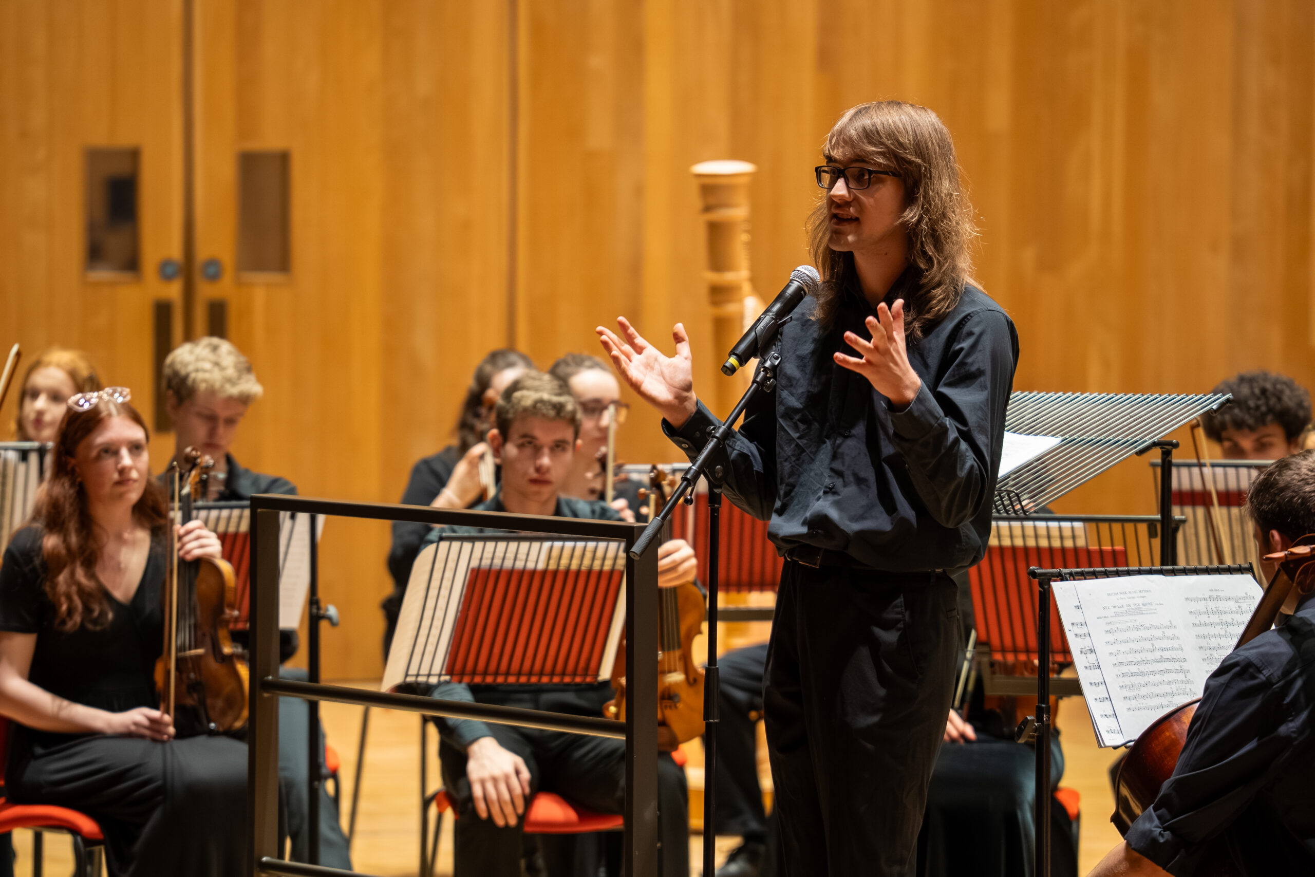Fern Warwick, wearing black-rimmed spectacles, long brown hair and black shirt, is talking to the orchestra.