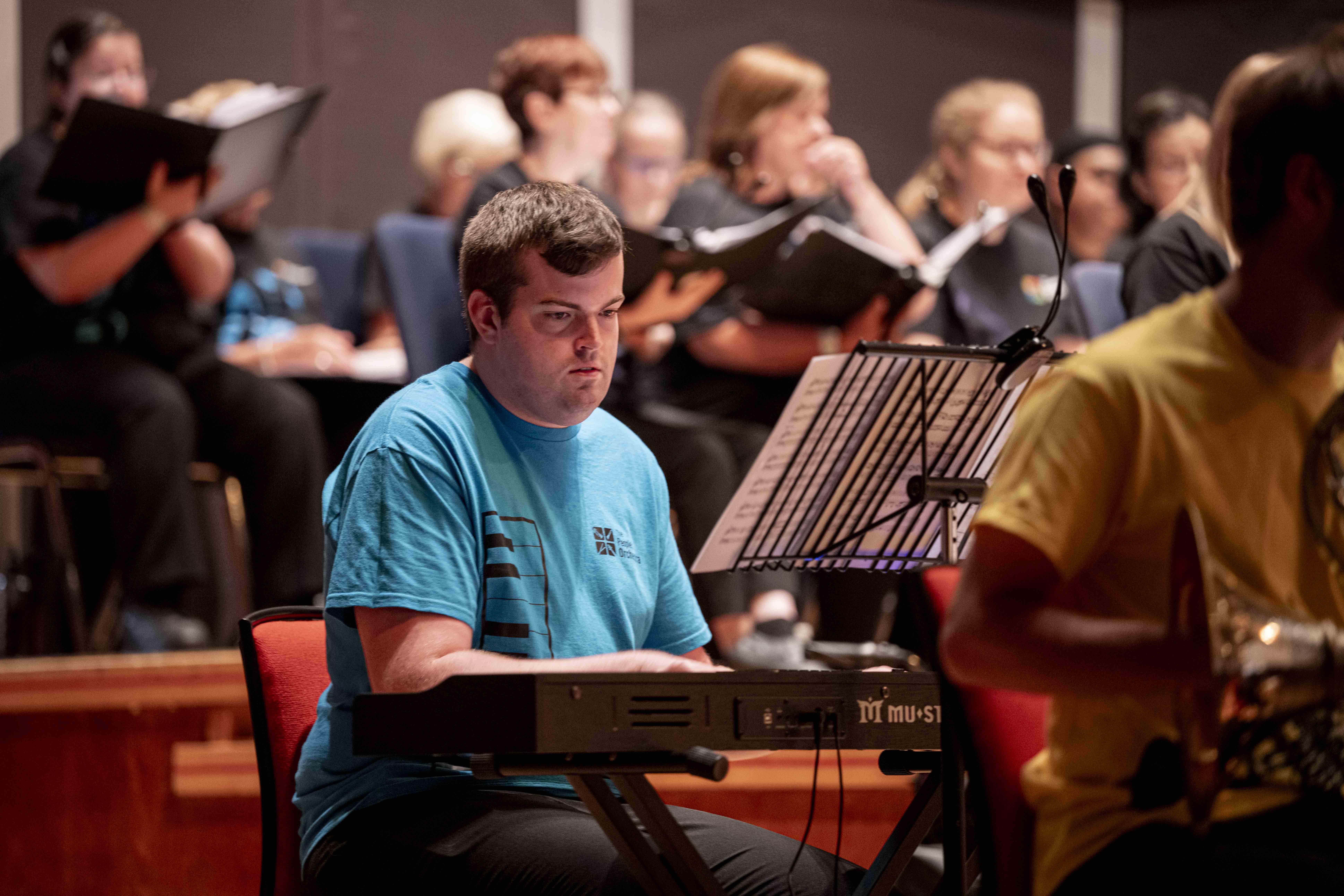A young man with brown hair plays the piano at Birmingham Symphony Hall. He wears a blue TPO-branded t-shirt. 