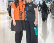 Two women are smiling, holding each other close. The woman on the left has black hair and is wearing an orange TPO-branded t-shirt. The other woman on the right has white hair and is wearing a TPSC branded t-shirt, black with a blue treble clef.